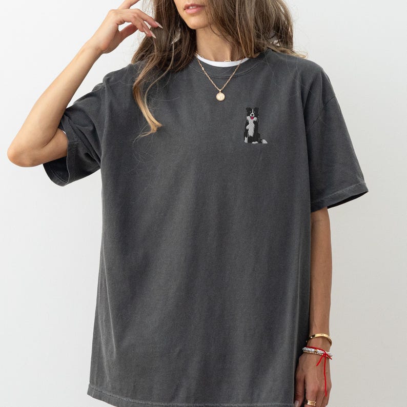 May include: Dark gray t-shirt with a small embroidered black and white dog design on the chest. The shirt has short sleeves and a relaxed fit. The model is wearing a white undershirt and gold jewelry.