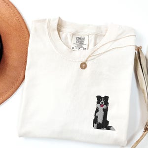 May include: Cream-colored t-shirt with a small embroidered black and white dog design. The dog has a red tongue. A gold necklace with a round pendant rests on the shirt. The shirt is folded and has a Comfort Colors tag.