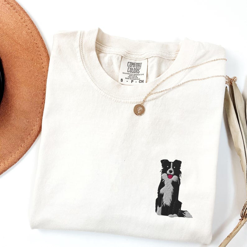 May include: Cream-colored t-shirt with a small embroidered black and white dog design. The dog has a red tongue. A gold necklace with a round pendant rests on the shirt. The shirt is folded and has a Comfort Colors tag.