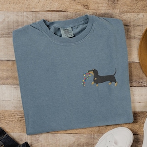 May include: A blue-grey t-shirt featuring an embroidered dachshund dog holding a string of colorful Christmas lights. The shirt is made of a soft material and has a classic crew neck design. The Comfort Colors brand tag is visible.