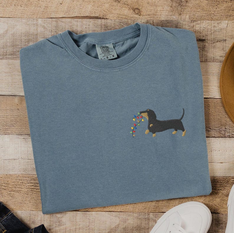 May include: A blue-grey t-shirt featuring an embroidered dachshund dog holding a string of colorful Christmas lights. The shirt is made of a soft material and has a classic crew neck design. The Comfort Colors brand tag is visible.