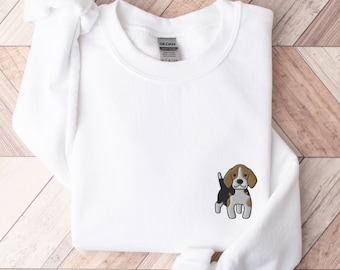 May include: White sweatshirt with a small embroidered beagle dog on the front. The beagle has brown, black, and white markings. The sweatshirt is folded neatly on a light wood surface.