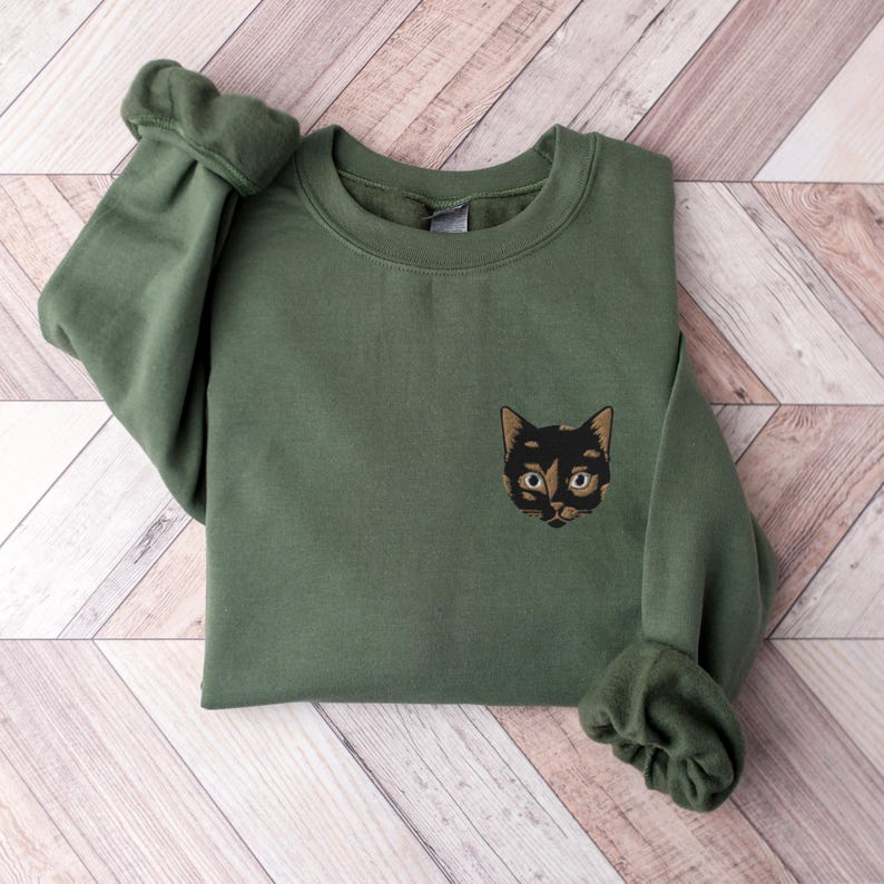 Op de afbeelding: Een groen sweatshirt met een geborduurd zwart en bruin kattengezicht op de voorkant.