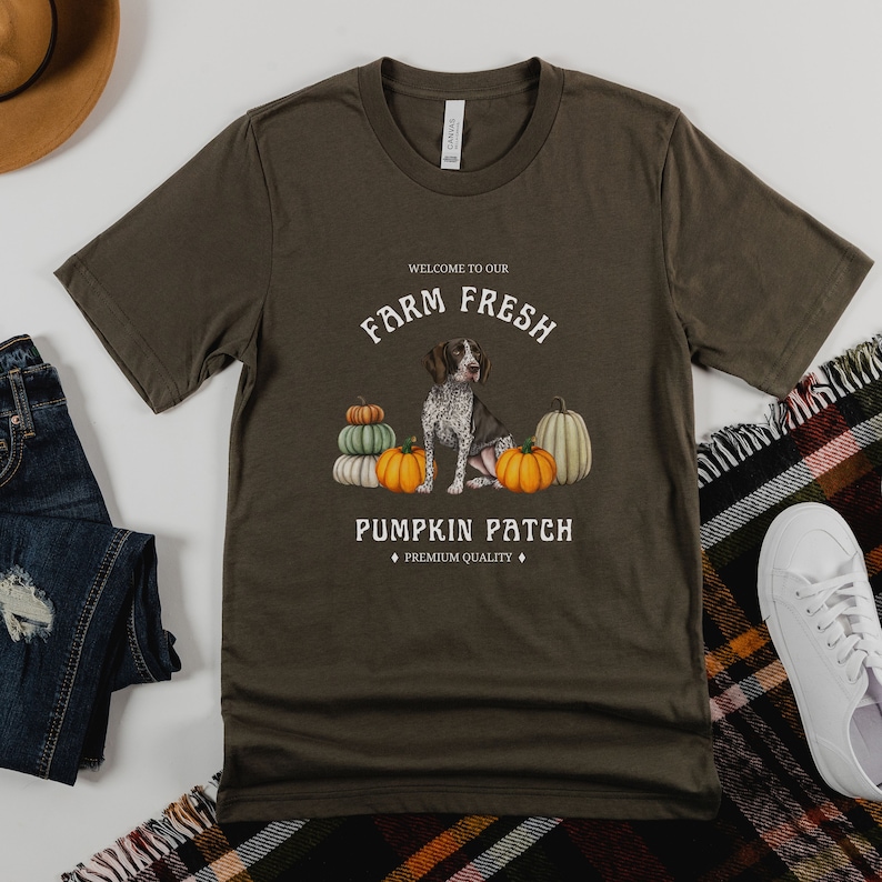 May include: A green t-shirt with a white graphic design featuring a dog sitting amongst pumpkins. The text on the shirt reads "Welcome to our Farm Fresh Pumpkin Patch, Premium Quality".