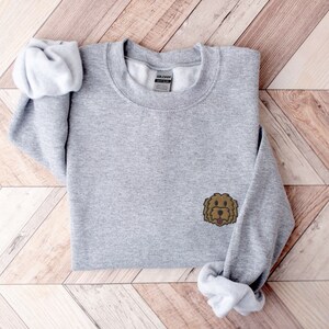 May include: A gray sweatshirt with a brown doodle of a dog's face on the left side of the chest. The sweatshirt is folded and rolled up on a wooden surface.