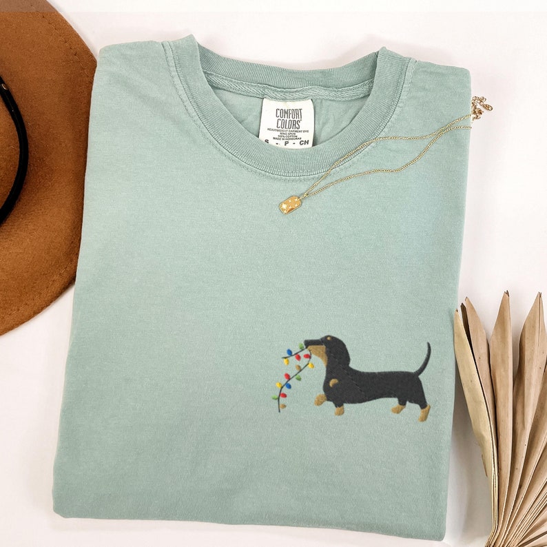May include: A light green t-shirt with a dachshund dog embroidered on the front, holding a string of colorful Christmas lights. A gold necklace and a brown hat are also visible.