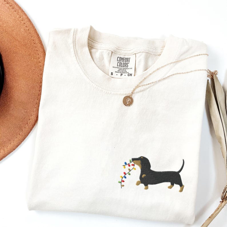May include: Cream-colored t-shirt with a dachshund dog graphic carrying a string of colorful Christmas lights. The shirt has a crew neck and short sleeves. A gold necklace with a pendant is draped across the shirt.