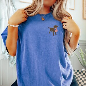 Puede incluir: Una camiseta azul con una silueta negra de un perro sosteniendo una bandera estadounidense en su boca.