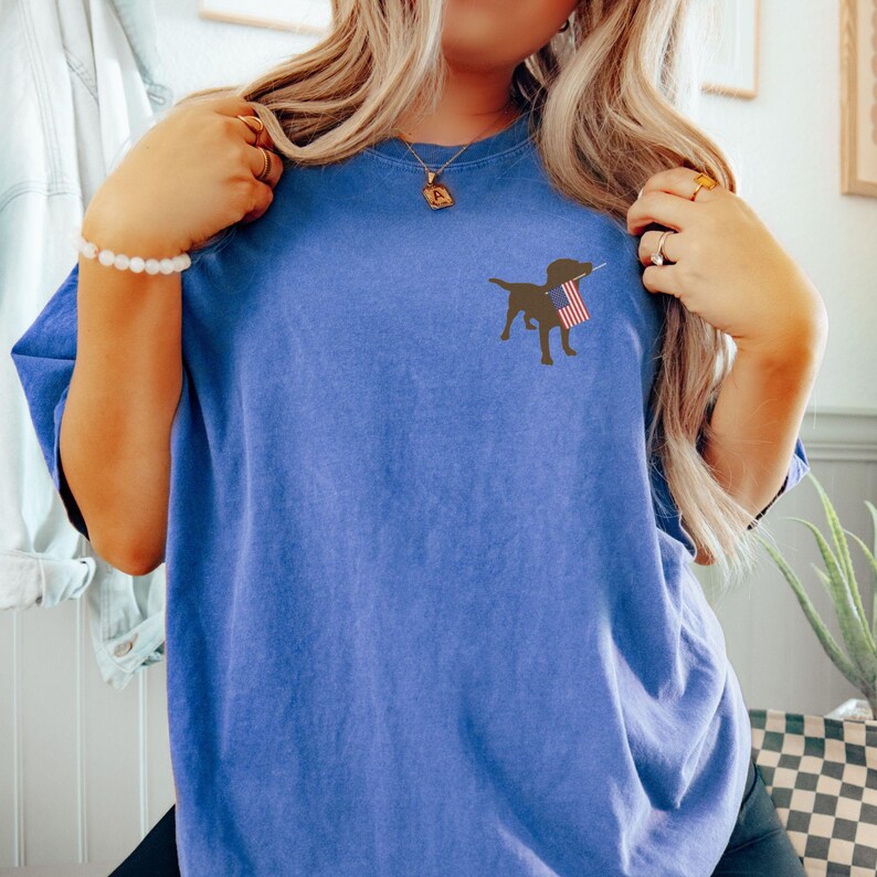 Puede incluir: Una camiseta azul con una silueta negra de un perro sosteniendo una bandera estadounidense en su boca.