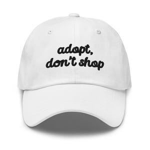 May include: White baseball cap with the embroidered text "adopt, don't shop" in black cursive font. The cap has a curved brim and a button on top. A casual accessory for everyday wear.