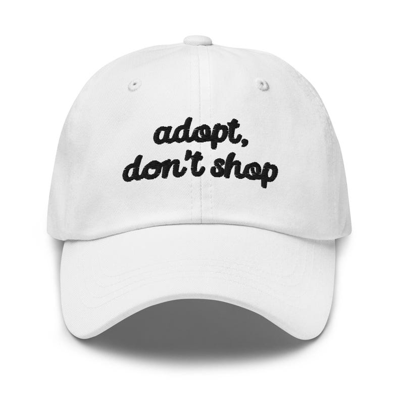 May include: White baseball cap with the embroidered text "adopt, don't shop" in black cursive font. The cap has a curved brim and a button on top. A casual accessory for everyday wear.