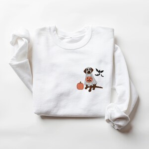 May include: White sweatshirt featuring a brown dog dressed as a ghost, holding a jack-o'-lantern. The dog is next to a pumpkin, with bats flying in the background.