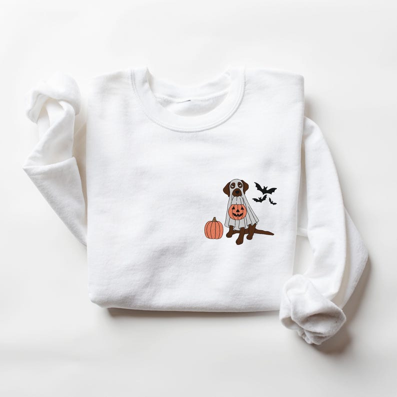 May include: White sweatshirt featuring a brown dog dressed as a ghost, holding a jack-o'-lantern. The dog is next to a pumpkin, with bats flying in the background.