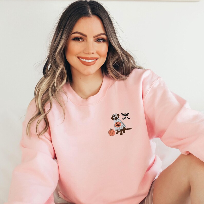 May include: A pink sweatshirt featuring a cute ghost dog wearing a pumpkin bucket and bats flying around it. The dog is brown and white.