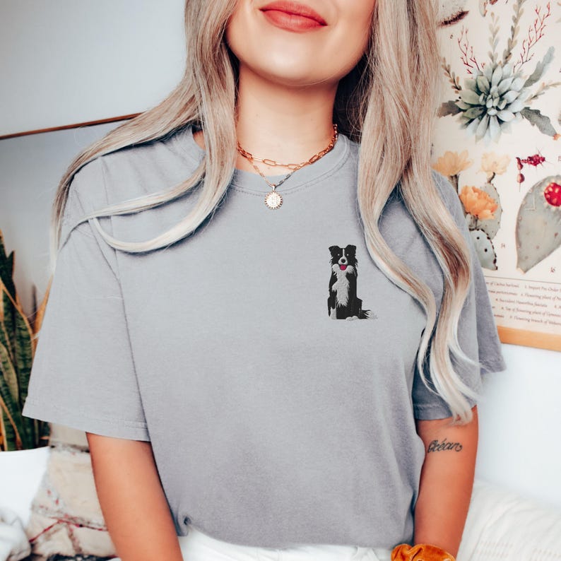 May include: A light gray t-shirt featuring a small embroidered design of a black and white Border Collie dog with a red tongue. The dog is sitting upright. The shirt has short sleeves and a crew neck.