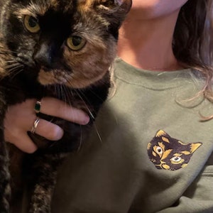 Op de afbeelding: Een persoon die een schildpadkat vasthoudt. De persoon draagt een groen sweatshirt met een geborduurd kattengezicht op de voorkant.