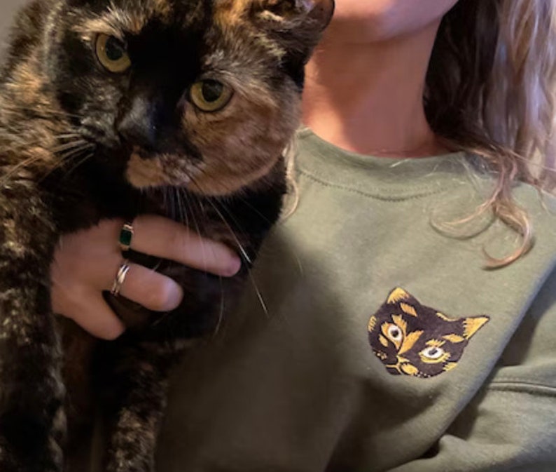 Op de afbeelding: Een persoon die een schildpadkat vasthoudt. De persoon draagt een groen sweatshirt met een geborduurd kattengezicht op de voorkant.