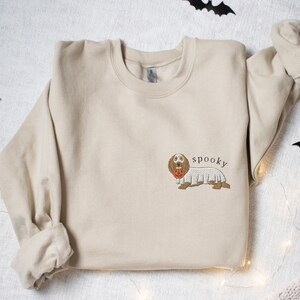 May include: A beige sweatshirt with a brown dachshund wearing a ghost costume and holding a jack-o'-lantern. The text "spooky" is printed above the dog.