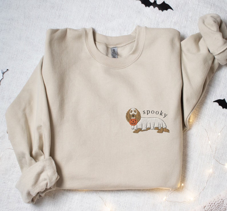 May include: A beige sweatshirt with a brown dachshund wearing a ghost costume and holding a jack-o'-lantern. The text "spooky" is printed above the dog.
