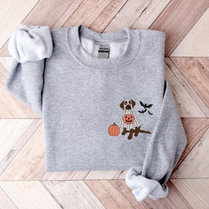 May include: A grey sweatshirt featuring a brown dog dressed in a white ghost costume, holding a jack-o'-lantern. The dog is standing next to a pumpkin. Three black bats are flying above the dog.