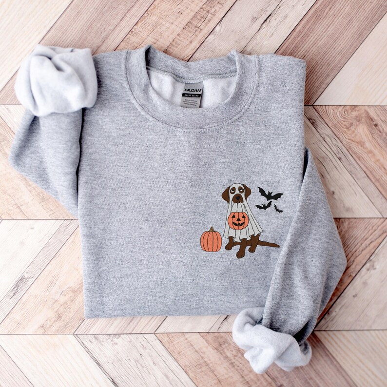 May include: A grey sweatshirt featuring a brown dog dressed in a white ghost costume, holding a jack-o'-lantern. The dog is standing next to a pumpkin. Three black bats are flying above the dog.