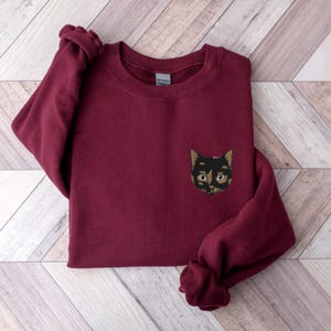 Op de afbeelding: Een bordeauxrood sweatshirt met een geborduurd kattengezicht in zwart en bruin op de linkerborst.