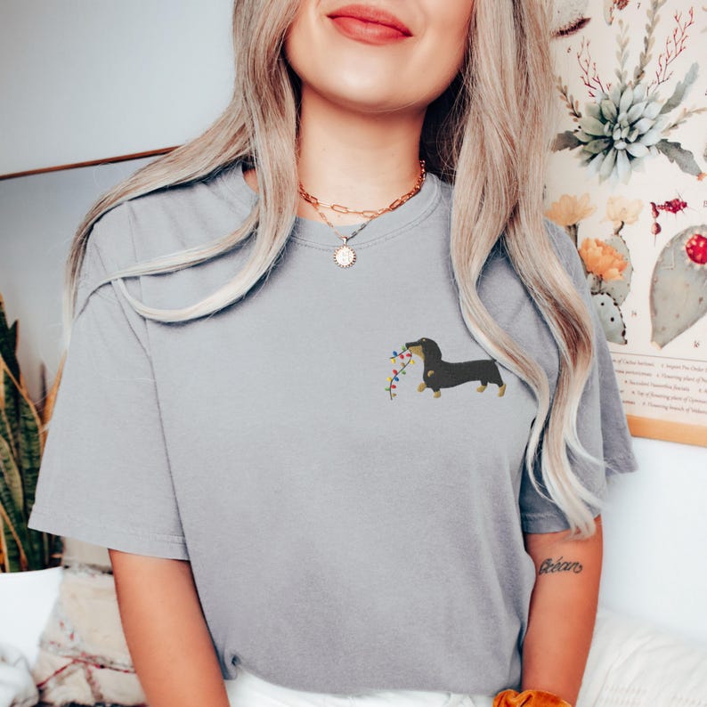 May include: A light gray t-shirt featuring a dachshund dog carrying a string of colorful lights. The dog is black and brown. The shirt has short sleeves and a relaxed fit.