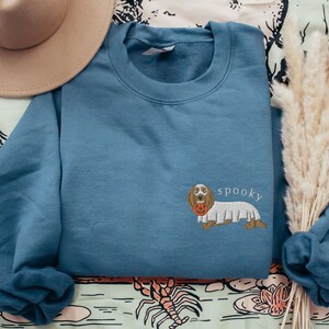 May include: A blue sweatshirt with a white ghost costume dachshund holding a pumpkin. The text "spooky" is embroidered on the sweatshirt.