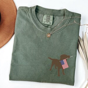 Puede incluir: Una camiseta verde con una silueta marr&oacute;n de un perro sosteniendo una bandera estadounidense. La camiseta tiene una etiqueta que dice "Comfort Colors" y "S - P - CH".