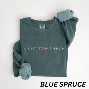 May include: Blue spruce sweatshirt featuring the text "ADOPT DON'T SHOP" in white and a small red heart. The sweatshirt has a crew neck and long sleeves with cuffed ends. The words "BLUE SPRUCE" are printed at the bottom.