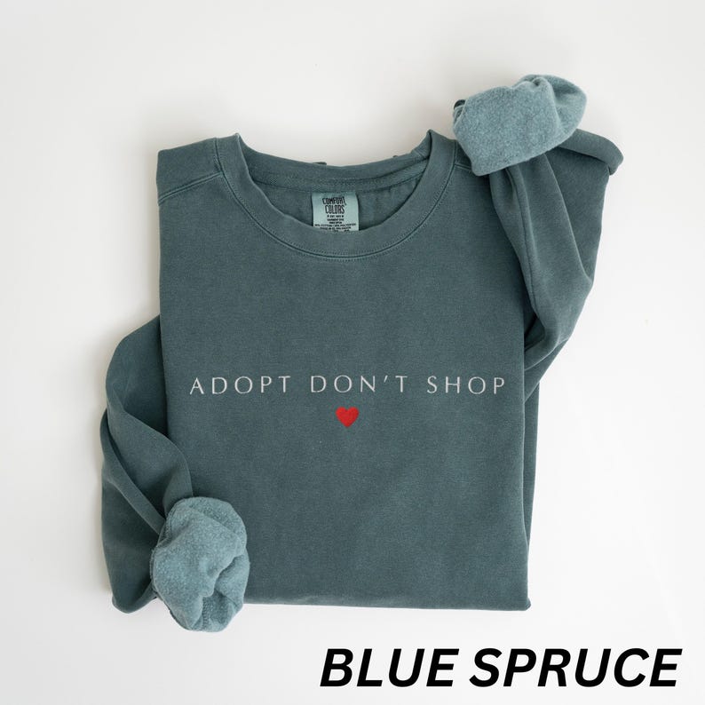 May include: Blue spruce sweatshirt featuring the text "ADOPT DON'T SHOP" in white and a small red heart. The sweatshirt has a crew neck and long sleeves with cuffed ends. The words "BLUE SPRUCE" are printed at the bottom.