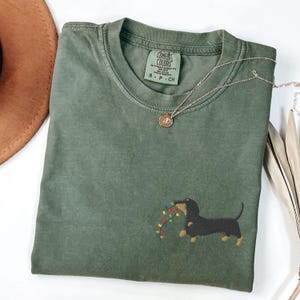 May include: An olive green t-shirt with a dachshund dog embroidered on the front, carrying a string of colorful Christmas lights. The shirt has a crew neck and short sleeves. A gold necklace with a pendant is draped across the shirt.