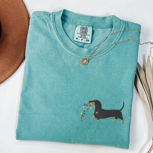 May include: A teal Comfort Colors t-shirt with a dachshund dog embroidered on the front, holding a string of colorful Christmas lights. A gold necklace with a pendant is draped across the shirt.
