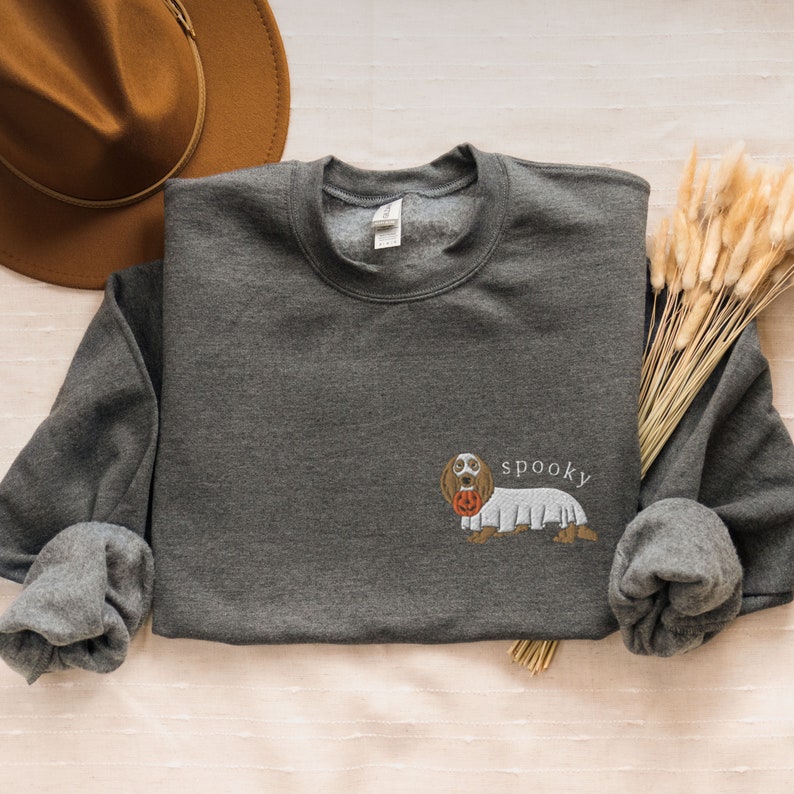 May include: A dark gray sweatshirt with a white embroidered image of a dachshund wearing a ghost costume and holding a pumpkin. The text "spooky" is embroidered above the dog.