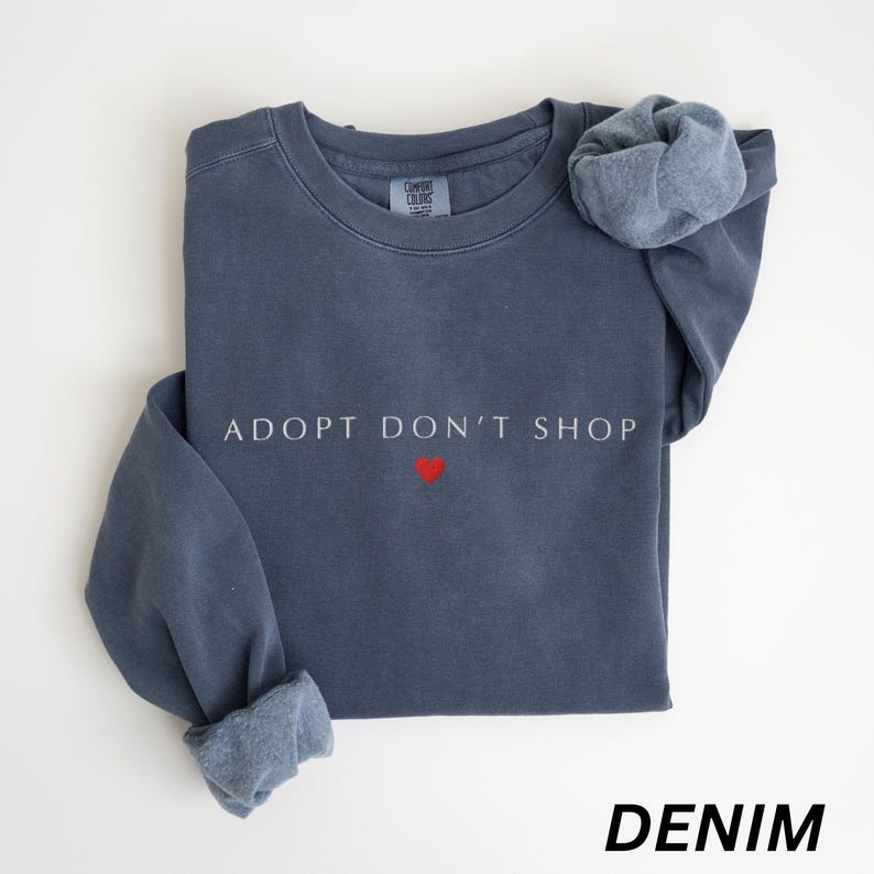 May include: A denim blue sweatshirt with the text "ADOPT DON'T SHOP" in white, and a small red heart. The sleeves are cuffed, and the word "DENIM" is printed at the bottom. The sweatshirt is made of a soft material.