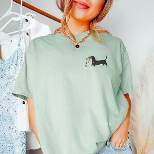 May include: A light green t-shirt featuring a dachshund dog carrying a string of colorful lights. The dog is black with tan legs and ears. The shirt is a relaxed fit and has short sleeves.