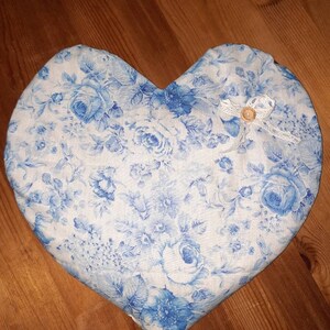 Könnte beinhalten: Ein herzförmiges Kissen mit weißem Hintergrund und einem blauen Blumenmuster. Das Kissen hat einen kleinen weißen Knopf mit braunem Kern.