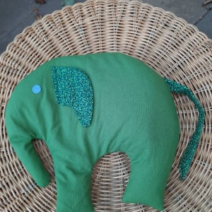 Könnte beinhalten: Ein grünes, elefantenförmiges Kissen mit einem gemusterten Ohr und Schwanz. Das Kissen hat ein kleines blaues Kreisdetail und ist aus weichem Stoff gefertigt. Das Elefantenkissen ist ein dekorativer Artikel.