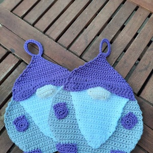 Könnte beinhalten: Zwei gehäkelte Topflappen in Form von Gnomen mit violetten Mützen und weißen Bärten. Die Gnome sitzen auf einer Holzoberfläche.
