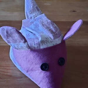 Könnte beinhalten: Eine lila Stoffmaus mit einem weiß-rosa gemusterten Kopf und schwarzen Knopfaugen.