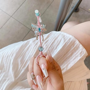 May include: A pink and silver wire wrapped ring with a blue butterfly charm and a clear crystal bead. The ring is on a finger with long, clear, iridescent nails.