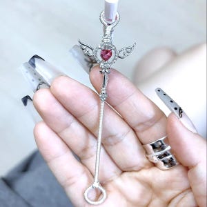 May include: A silver wand-shaped accessory featuring a pink heart-shaped gem, small wings, and a long, slender stem. The wand has a ring at the base and decorative detailing at the top. The design is intricate and detailed.