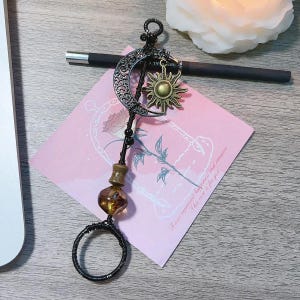 May include: A black metal sun and moon charm with a brown glass bead and a black wire ring. The charm is hanging from a pink card with the text "Roses represent hope, love and promise. This rose is for you."