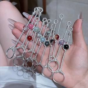 Op de afbeelding: Een verzameling zilverkleurige metalen ringhouders met decoratieve edelsteenaccenten. Elke houder heeft een anders gekleurde edelsteen, waaronder roze, rood, groen en zwart. De ringhouders worden in een hand gehouden.