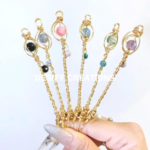 Könnte beinhalten: Sechs goldfarbene, drahtgewickelte Zauberstäbe mit verschiedenen farbigen Glasperlen. Die Perlen sind rosa, blau, grün, lila, schwarz und weiß.