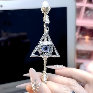 Op de afbeelding: Zilverkleurige hanger met een oog in een driehoek, een symbool dat vaak geassocieerd wordt met de Illuminati. De hanger heeft een blauw oog en hangt aan een gedraaide draad en een zilverkleurige bovenkant.
