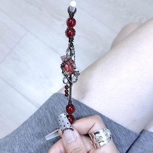 May include: A handmade hair stick with a dark wire design. The stick features red beads and a central decorative element with a red gemstone and floral accents. The hair accessory is held in a hand with a silver ring.