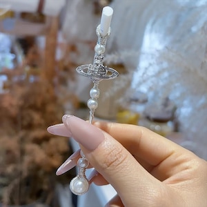 May include: A silver-coloured straw topper featuring a planet-shaped charm, embellished with clear crystals and faux pearls. The straw topper is designed to fit a standard straw. The design is elegant and decorative.