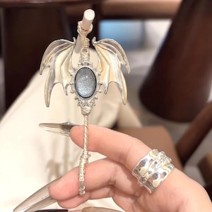 May include: A silver-coloured, dragon-winged accessory with an oval, glittery centre stone. The accessory has a detailed, twisted handle and is held near a silver ring with a textured design.