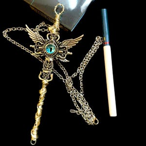 May include: A gold-toned necklace with a large pendant featuring a winged eye design. The pendant is detailed with intricate carvings and a blue gemstone. The necklace is paired with a silver chain.